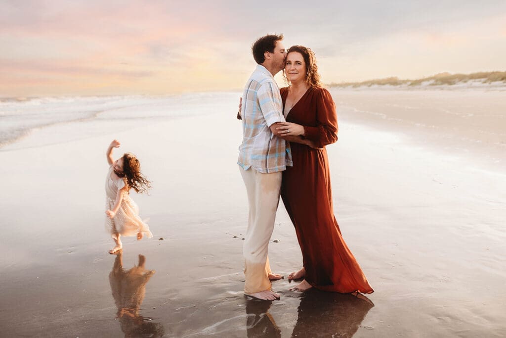 Parents embrace while their daughter runs along the beach during Family Photos on Kiawah Island with Holy City Family Photography after discovering the blog post about Things to Do on Kiawah Island with Kids.